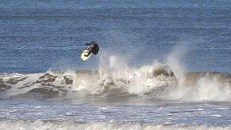 male advanced level surfer in black hooded wetsuit on a mid-height wave and completing an aerial manoeuvre, in cold water conditions on a bright and sunny day.