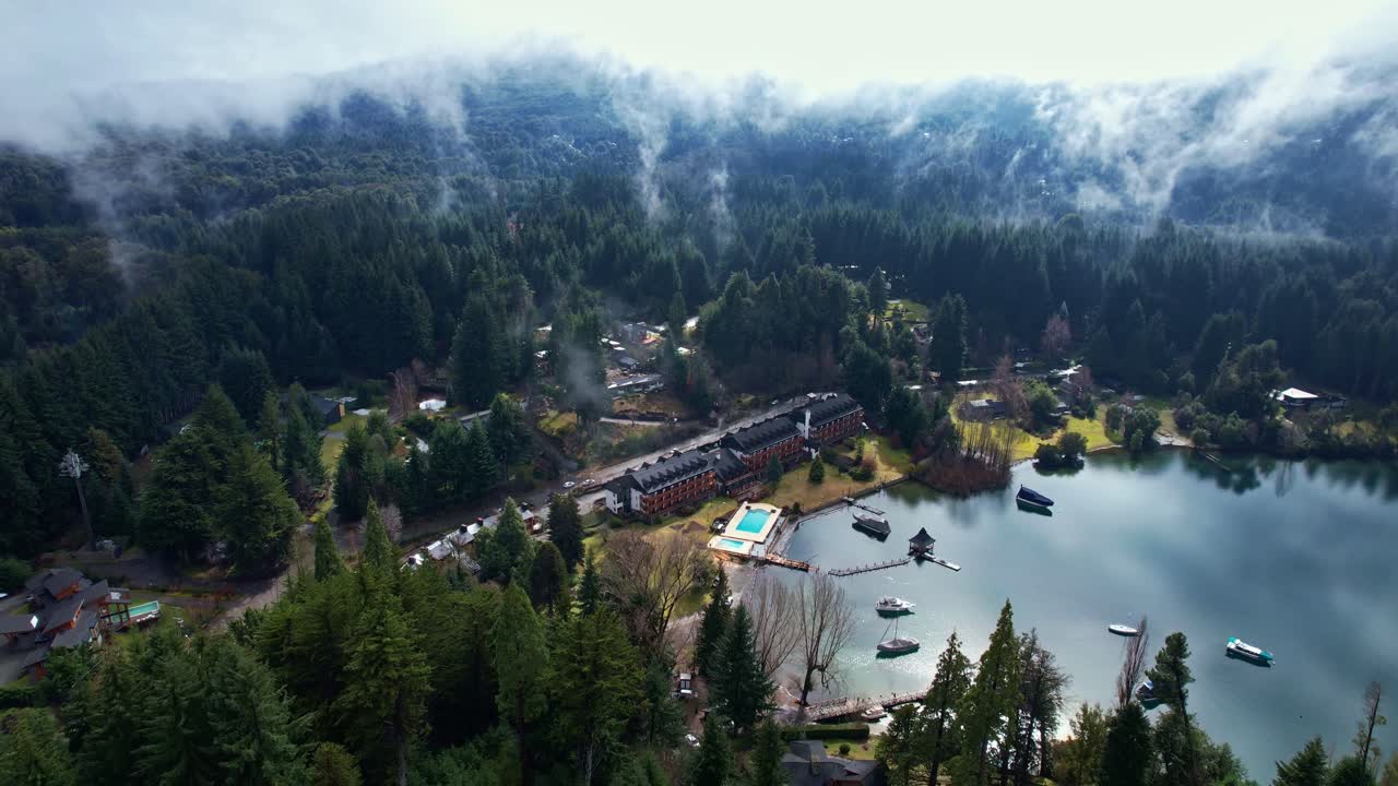 Drone flyver baglæns fra et luksuriøst bjergresort og en bugt ved Nahuel Huapi-søen. Det perfekt reflekterende vand trækker sig tilbage for at afsløre hvirvlende lavthængende skyer og tåge blandt tætte fyrreskove i Patagonien.
