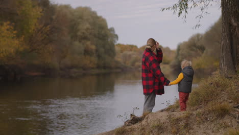 Una-Mujer-Y-Un-Niño-Pequeño-Están-Parados-En-La-Costa-Del-Río-En-El-Fin-De-Semana-Familiar-Del-Día-De-Otoño-En-La-Naturaleza,-Madre-E-Hijo-Caminan-En-El-Bosque-En-Un-Momento-Tranquilo-Y-Feliz-Juntos