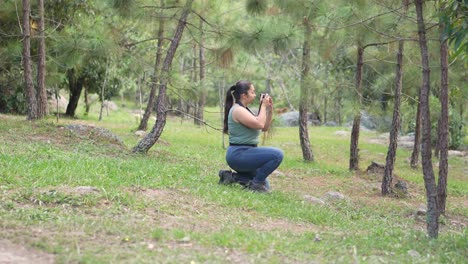 young female photographer crouches in the forest with a camera in her hands, taking pictures outdoors surrounded by nature