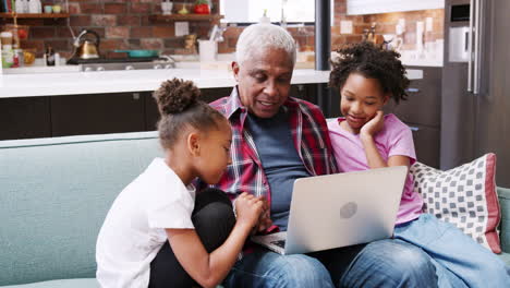 Abuelo-Sentado-En-El-Sofá-En-Casa-Con-Nietas-Usando-Una-Computadora-Portátil