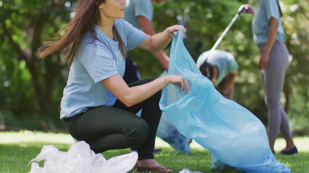 Donna caucasica sorridente e un gruppo eterogeneo di amici che raccolgono rifiuti in sacchi della spazzatura blu in una foresta.