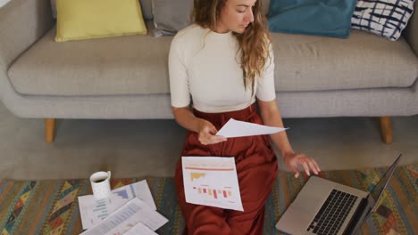 mujer caucásica trabajando en la sala de estar, sentada en el suelo con café y papeleo, usando computadora portátil