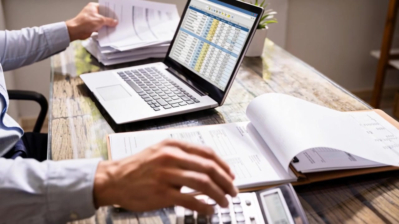 Noticing laptop spreadsheet worker in blue shirt typing using calculator reconciling papers at desk