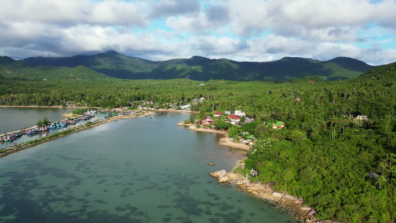 Aerial view of a coastal landscape featuring a harbor, palm trees, and mountains under a partly cloudy sky.