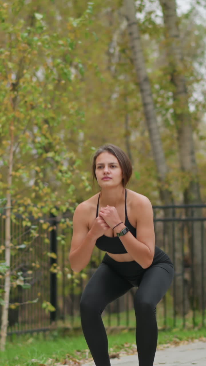 Athlete in black fitness wear performing squat exercise outdoors beside iron fence with dry trees in the background, focusing on fitness training, strength, and outdoor workout