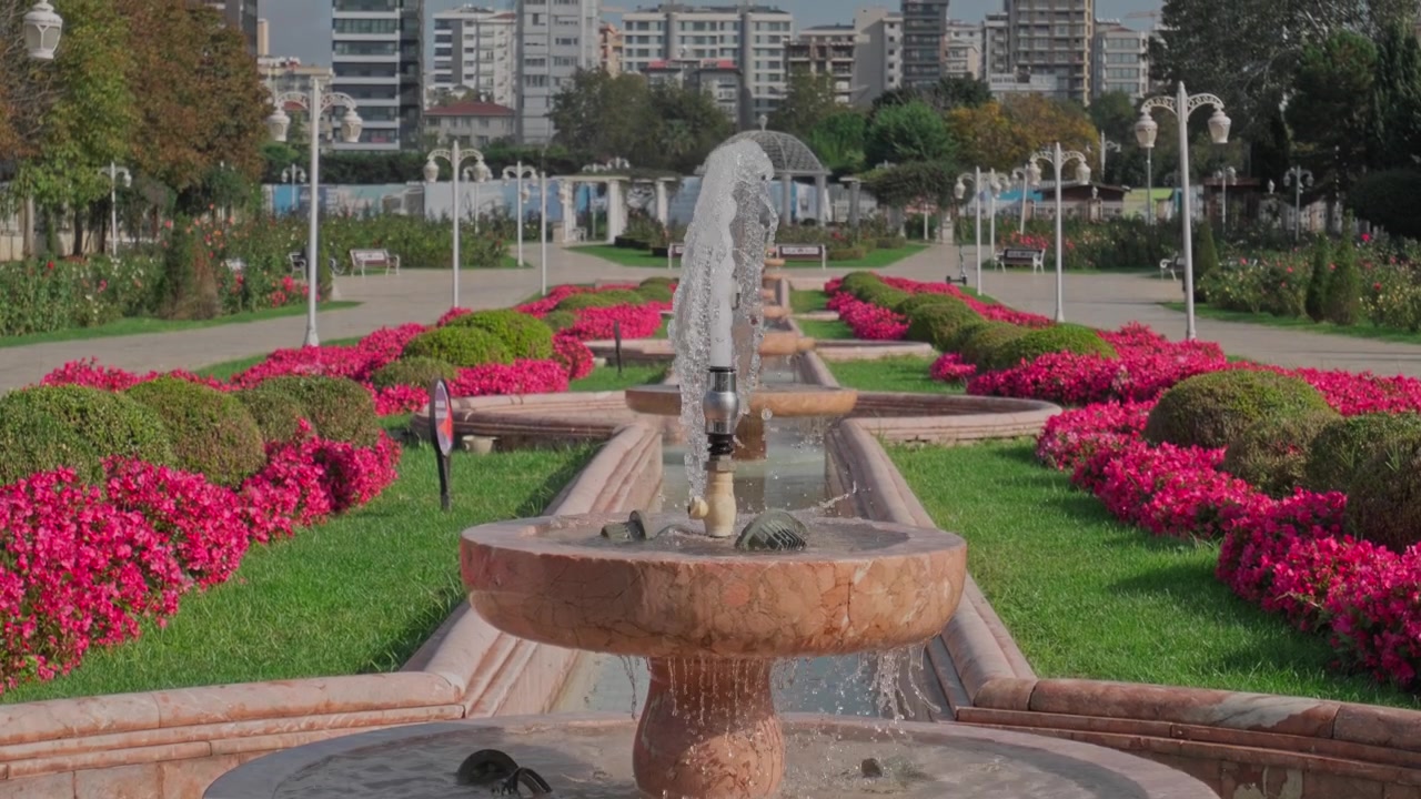 Vista de estilo aéreo de un jardín urbano formal que presenta una fuente de piedra central rodeada de parterres de flores rojos y rosas simétricos con césped verde en un espacio público urbano.