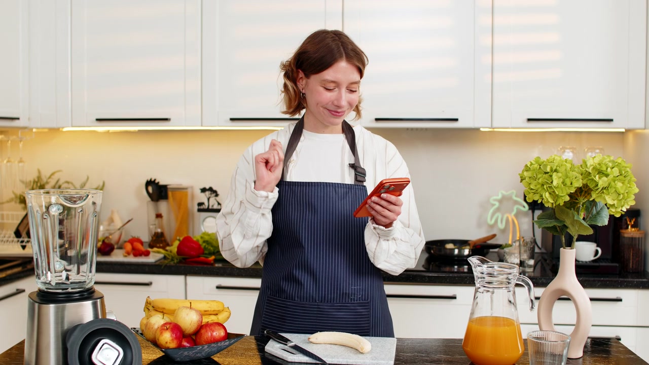 Giovane donna che controlla lo smartphone per la lista della spesa mentre prepara un frullato dietetico sano nella cucina di casa.
