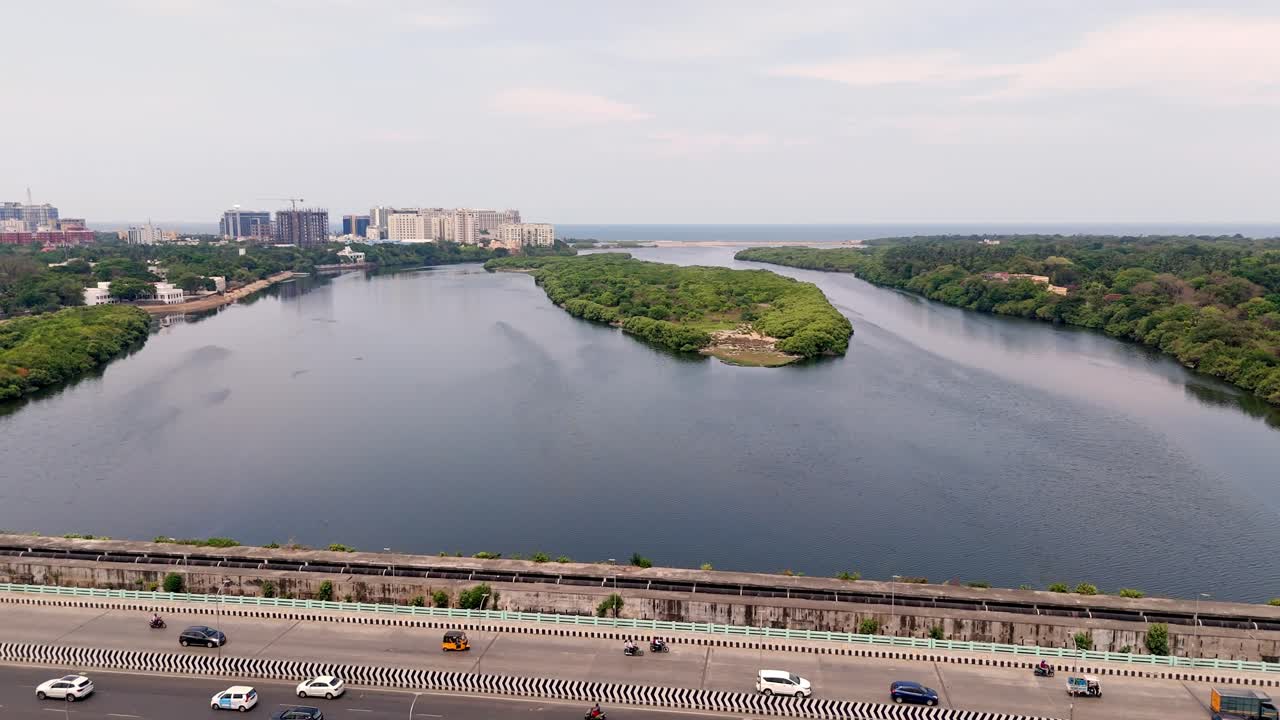 Aerial side view of vehicles apssing over bridge with small green lush island at background.