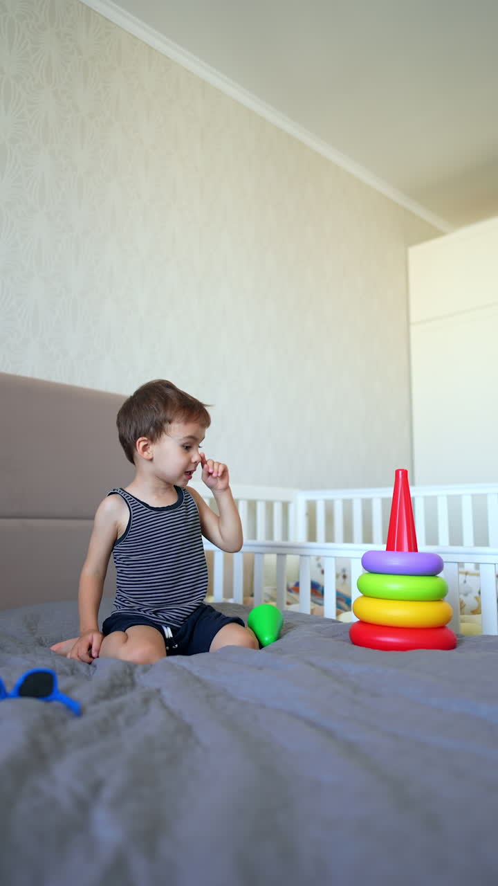 Cute Caucasian baby boy sits on the bed assembling a toy pyramid. Toddler kid plays at home cheerfully. Vertical video.