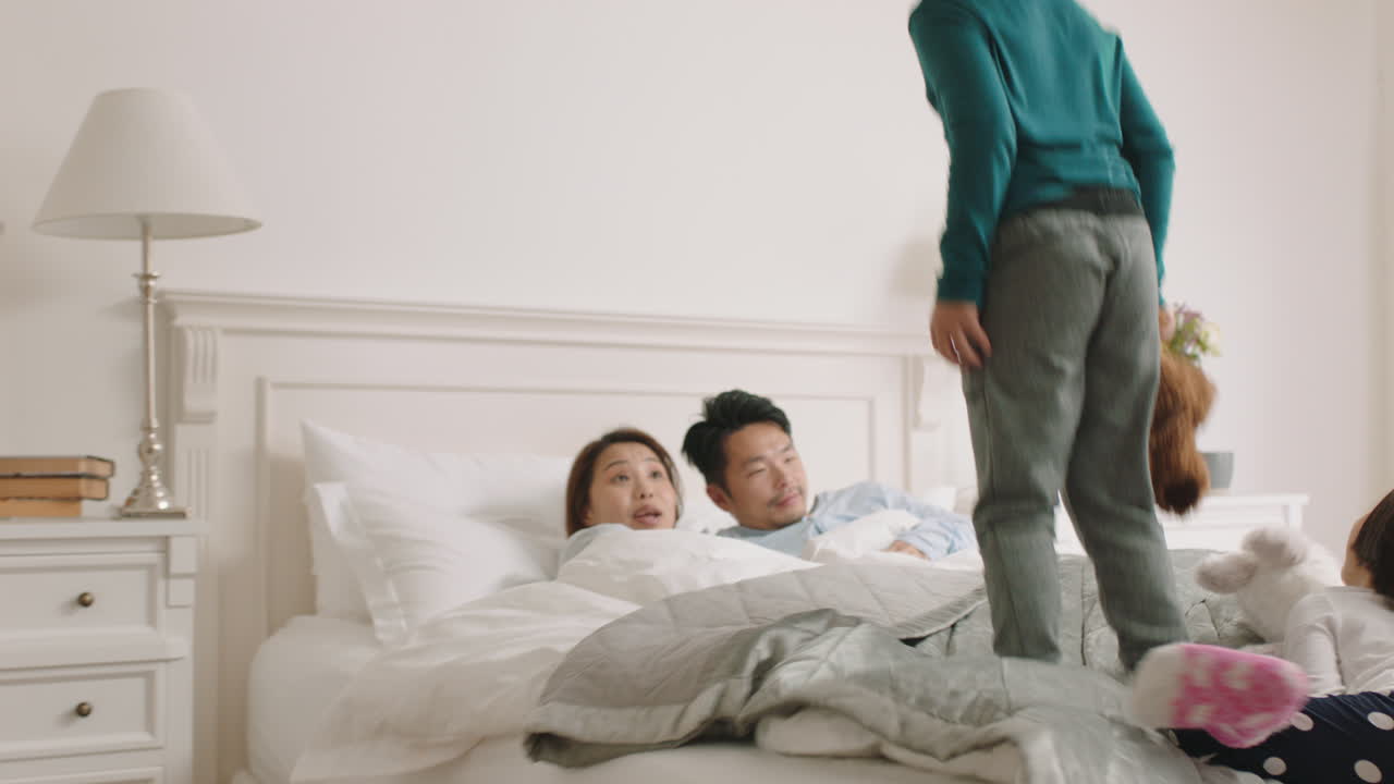 행복한 아시아 아이들이 침대에 뛰어들고 깨어나는 엄마와 아빠가 주말에 집에서 아이들과 함께 놀이하는 아침을 즐기고 있습니다. 4k 영상