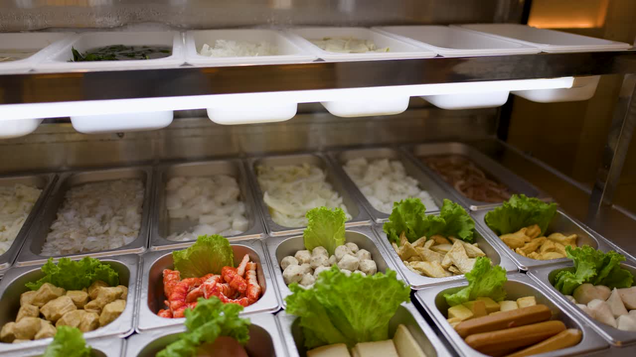 Camera pans across buffet trays of raw noodles, vegetables, and hotpot ingredients under bright lighting