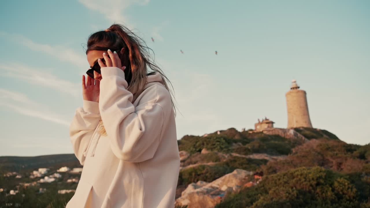 Ragazza moderna e alla moda con occhiali da sole che si gode il tramonto in spiaggia con un faro sullo sfondo