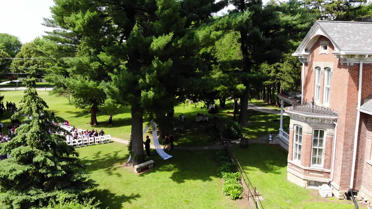 Aerial drone shot that begins on an old historical brick home and ends on a beautiful outdoor wedding with the seats starting to fill up.  This wedding is located in Nevada, Iowa.