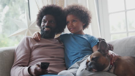 Smiling-middle-aged-black-father-and-pre-teen-son-reclining-on-sofa-together-watching-TV-with-their-pet-dog,-dad-holding-remote-control,-low-angle,-close-up