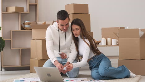 pareja joven en una casa nueva sentada en la alfombra con una laptop eligiendo colores para la decoración mientras se besa