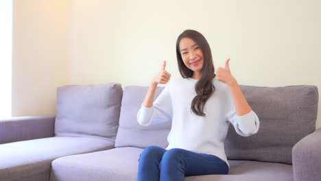 an asian thai woman showing big thumbs up gesture while sitting on the couch in casual clothes at home interior