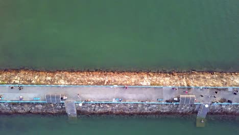 a dynamic top-down aerial shot of a breakwater jetty made out of stones with a paved pathway for civilians