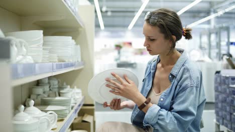 una chica en una tienda mira platos de cerámica blanca. gira el plato en sus manos, elige. mira los platos del estante inferior de la tienda. bajó. variedad de productos