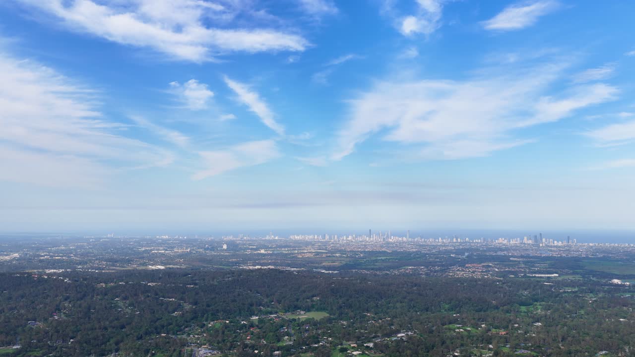 Drone pans above lush hinterland, revealing Gold Coast cityscape, skyscrapers, and coastal suburbs under sunlight