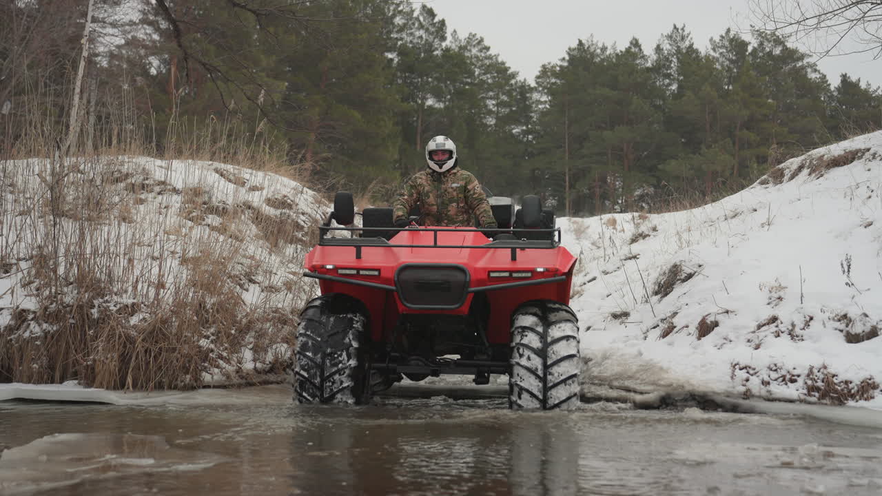 Quad rosso che attraversa un ruscello ghiacciato, il pilota negozia un guado poco profondo, schizzi d'acqua intorno ai pneumatici tassellati, cumuli di neve sulle rive, prospettiva frontale bassa, solchi compattati, luce invernale smorzata, passo calmo e costante
