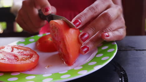 las manos femeninas usan un pequeño cuchillo para cortar tomate rojo maduro en el plato, de cerca