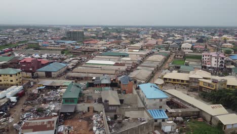 Aerial-view-small-community-in-Lagos-Nigeria-and-drone-moving-to-reveal-an-industry,-a-bus-park-and-a-bridge-with-cars-and-trucks-moving-speedily