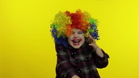 little child girl clown in colorful wig hides behind her hands and shows funny faces. halloween