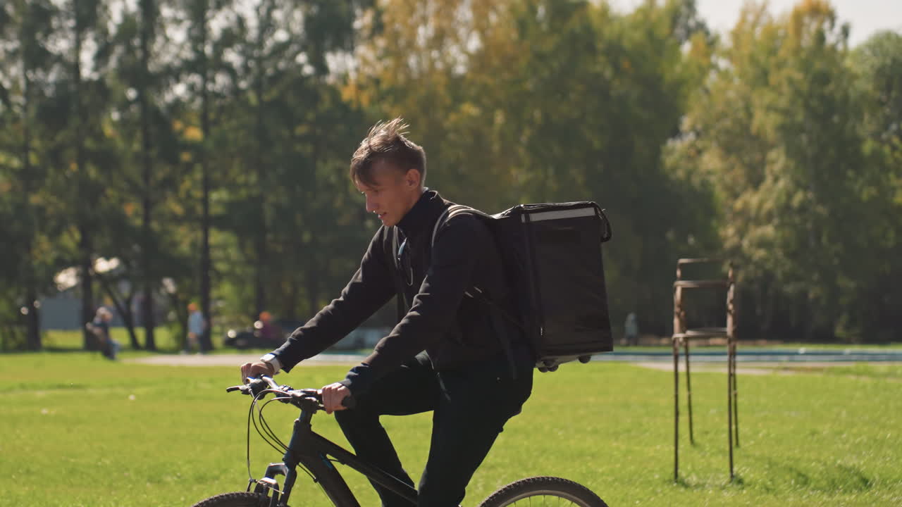caucasian courier cycling with delivery bag through sunny park grass, wearing black jacket, carrying insulated backpack, navigating paved path and sidewalk, checking smartphone app, steady pedal