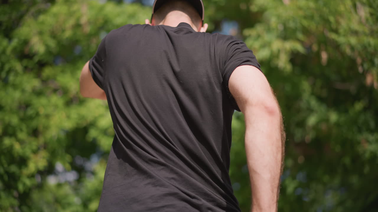 White Man Trainer Warming Up Outdoors Bending Into Stretch With Cap And Khaki Shorts, Tattoo And Beard Visible, Leafy Green Background, Focused Breathing And Dynamic Posture For Mobility Work