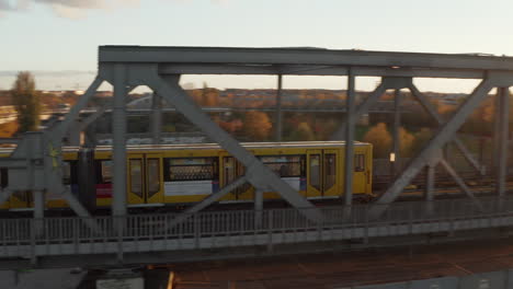 Yellow-Subway-Train-passing-bridge-through-urban-environment--in-Berlin,-Germany-in-beautiful-golden-hour-Sunset-light,-Aerial-tracking-follow-shot