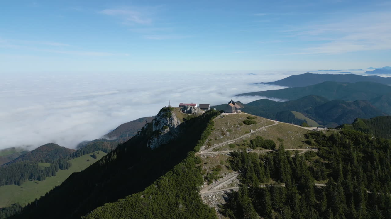 Breathtaking aerial shot of a remote mountaintop lodge perched on a ridge, surrounded by winding trails, dense green forests, and a vast blanket of clouds stretching across distant valleys.