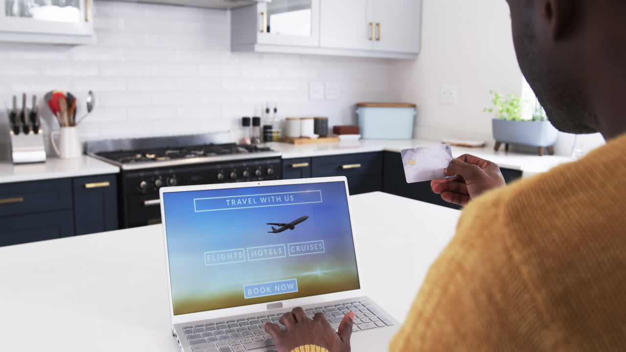 African American man booking holiday travel online using laptop in modern kitchen