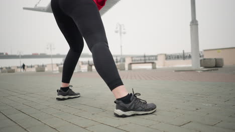 low-angle view of lady in legging stretching into a lunge during outdoor winter workout under modern canopy structure, with blurred pedestrian and distant bridge with cars in foggy urban surroundings