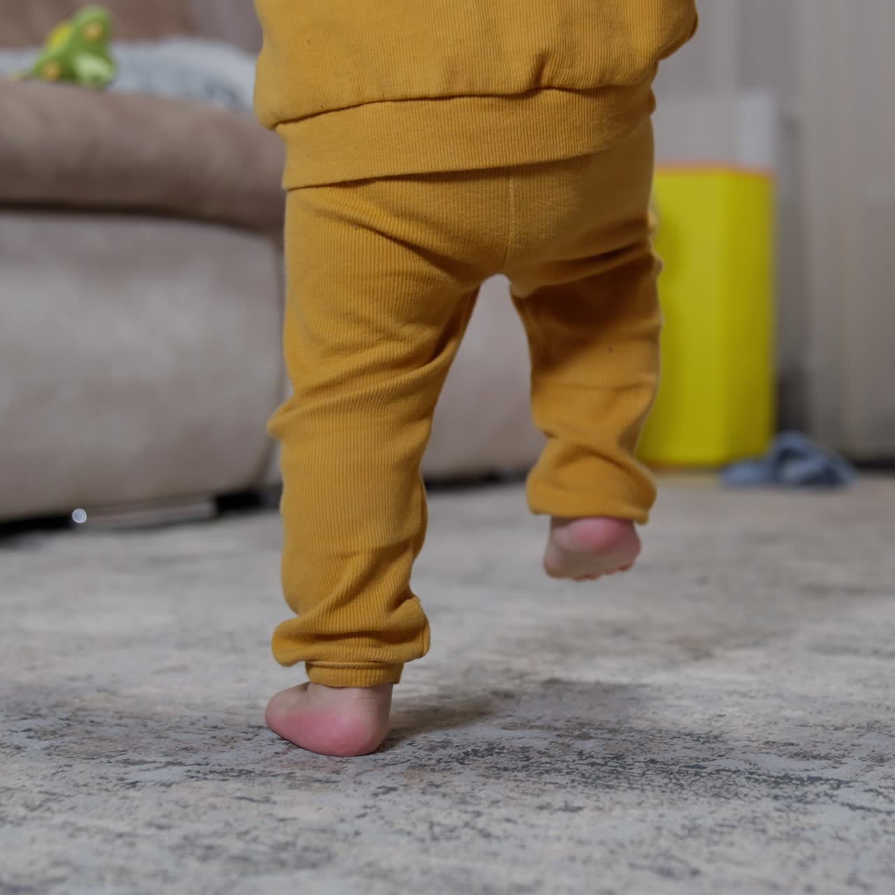 Child in orange sport suit is studying to walk indoors. Baby takes steps raising his feet high supported by his mom