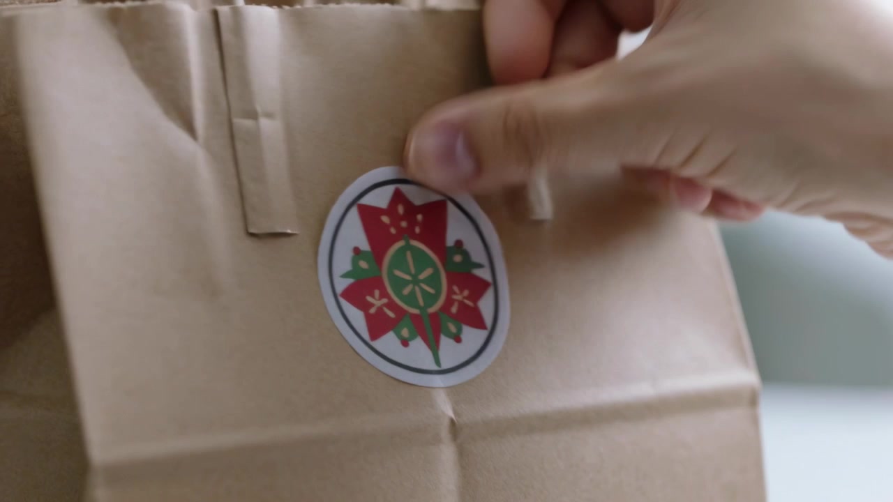 Mano destra che posiziona un adesivo floreale rotondo sulla parte anteriore di un sacchetto di carta marrone su un bancone, fissandolo