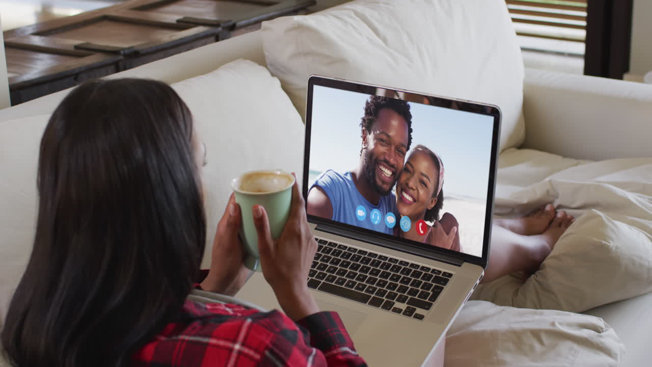 African american woman holding a coffee cup having a video call on laptop sitting on couch at home