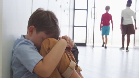 video de un triste niño caucásico sentado en el pasillo de la escuela, cubriendo la cabeza con las manos