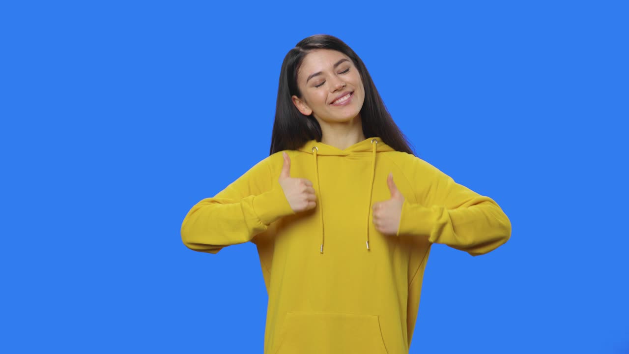 retrato de una bonita morena mostrando los pulgares hacia arriba, gesto como. mujer joven con cabello largo en sudadera amarilla posando en el estudio con fondo de pantalla azul. primer plano. cámara lenta lista 59.94fps