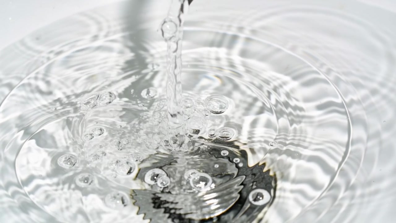 澄んだ水が浅い白い洗面器に流れ落ち、排水口の上に泡と波紋を作り出す様子