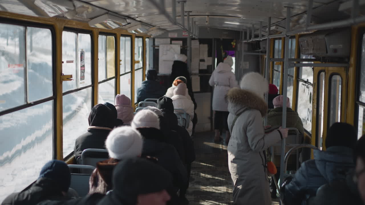 high angle interior view of crowded train during winter commute as passengers sit while others stand holding pole, carriage slows approaching stop, cold light through windows, urban transit mood