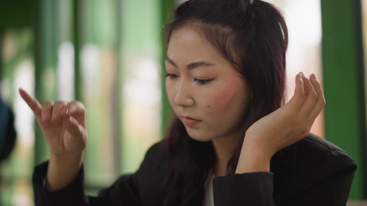 Cận cảnh người phụ nữ trẻ tựa đầu vào tay trong tư thế trầm tư, mặc áo blazer đen. Ánh sáng tự nhiên dịu nhẹ chiếu sáng khung cảnh với nền xanh lá và các chi tiết nội thất mờ ảo.