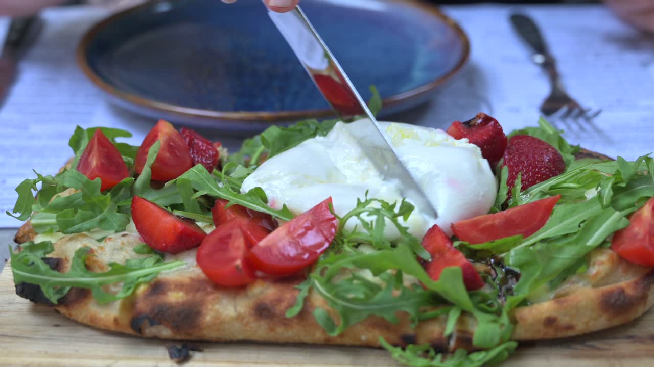 Woman cutting burrata on top of a pinsa with rucola and tomatoes