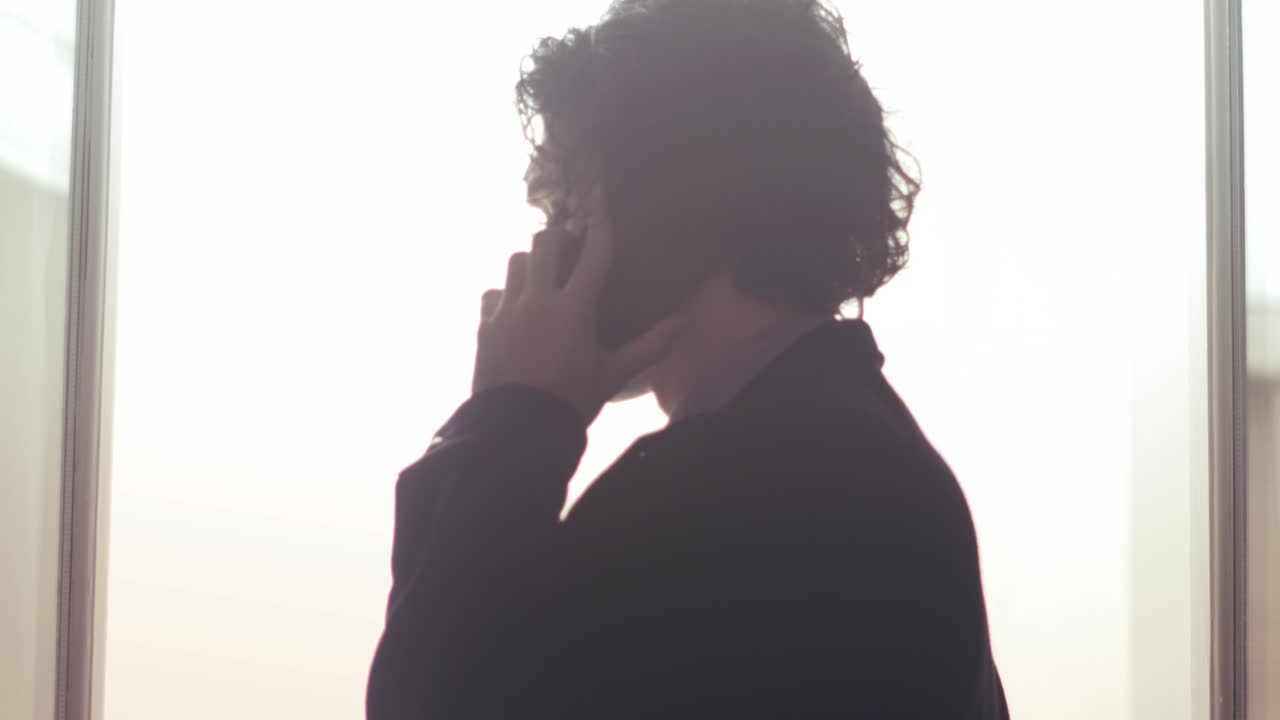 Silhueta de homem falando ao telefone junto à janela com luz mínima e brilhante
