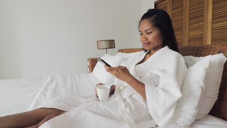 adult woman drinking coffee and using her mobile phone, browsin the internet while laying on a white bed wearing a white dressing gown