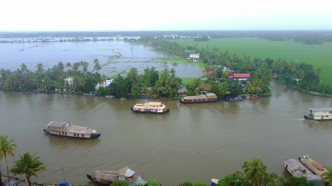 Scenic aerial view of houseboats cruising the wide, muddy backwaters of Kerala, surrounded by lush palm trees, village houses, and vast flooded paddy fields on an overcast day