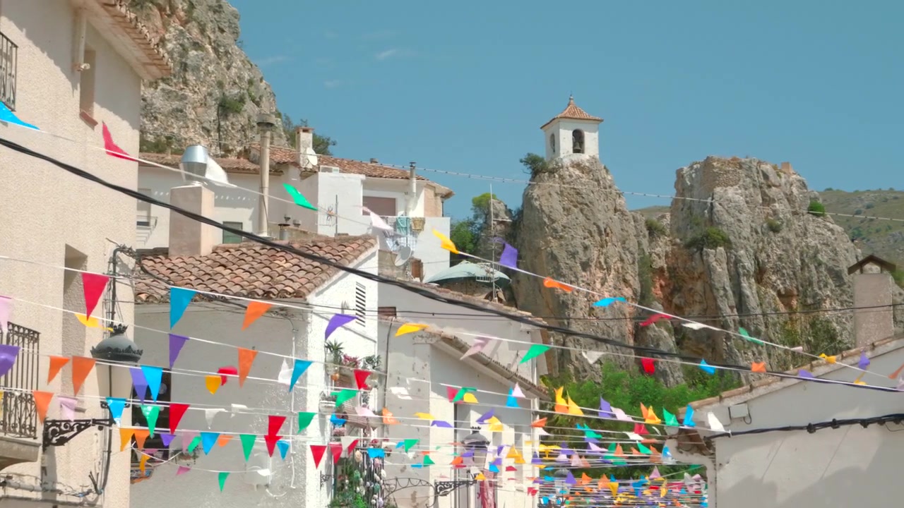 Luminosa strada di paese sotto un cielo azzurro limpido, con case intonacate di bianco, tetti in tegole di argilla, uno sfondo di scogliere rocciose, una piccola cappella in cima a una collina e molte bandierine colorate che creano un'atmosfera locale festosa.