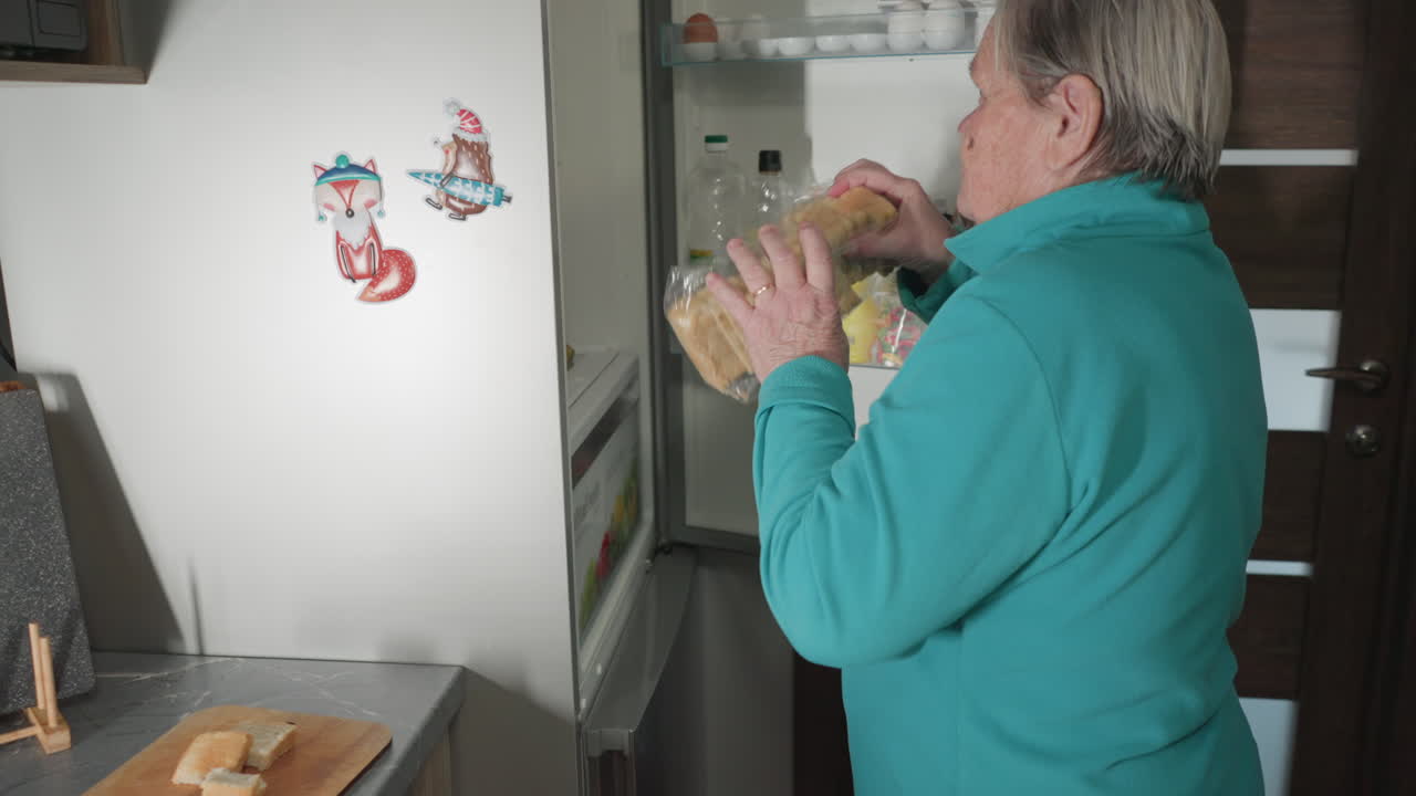 Person in green placing sliced bread back in its plastic bag after preparing slices on wooden cutting board, organizing bread in kitchen. Refrigerator and kitchen counter in background
