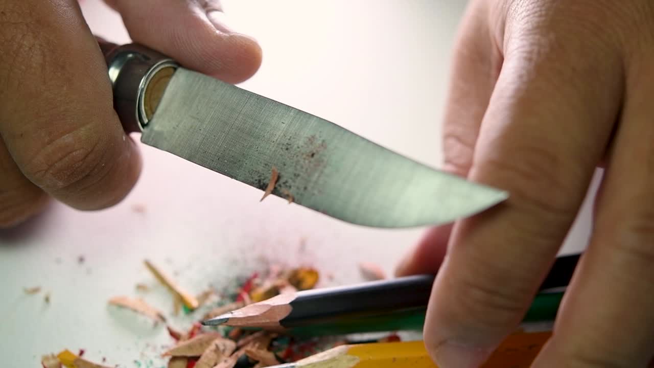Footage of hands slowly sharpening a pencil and some coloured pencils with a sharp knife