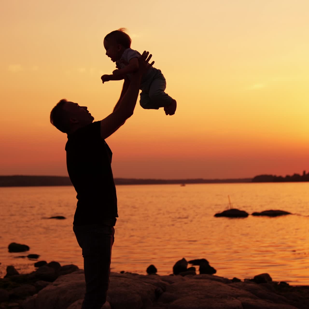 Un papà premuroso gioca con il suo bambino sul lungofiume. Un padre affettuoso lancia in aria suo figlio che gioisce. Il sole che tramonta sull'acqua sullo sfondo